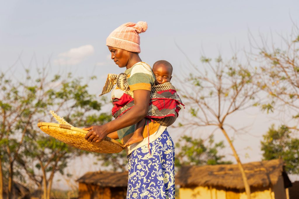 African mother carries baby on her back, outdoors in Mangochi, Malawi, showcasing traditional life.