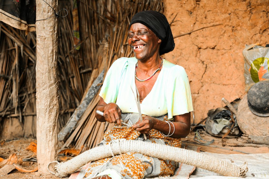 An African woman smiles as she weaves in a rural Tanzanian setting, showcasing traditional craftsmanship. The USAID freeze can easily wipe out that smile.